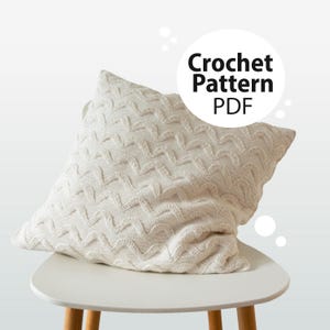 May include: A cream-colored, textured crochet pillow with a chevron pattern rests on a white chair. The pillow is square-shaped and appears soft. Text on the image reads "Crochet Pattern PDF".