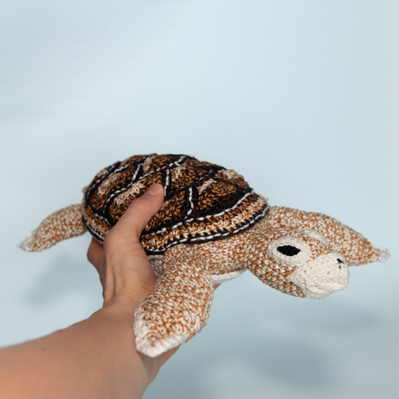 May include: A crocheted stuffed sea turtle toy with a brown and white shell and a beige body. The turtle has black eyes and a small, beige mouth.