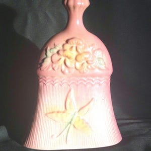 Könnte beinhalten: Eine rosafarbene Keramikglocke mit einem Blumendesign und einem Blatt auf der Unterseite. Die Glocke hat ein erhabenes Design aus Blumen und Blättern.