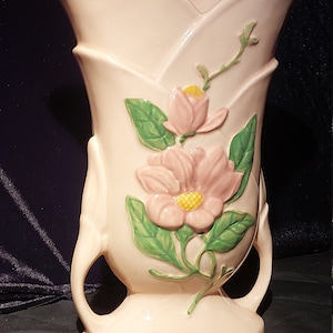 Può includere: Un vaso in ceramica vintage con una smaltatura rosa pallido e un motivo floreale. Il vaso ha due manici e un'ampia apertura. I fiori sono rosa con centri gialli e foglie verdi.