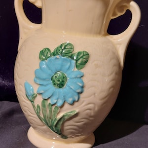 May include: A cream-colored ceramic vase with two handles and a blue and green floral design.