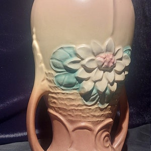 Op de afbeelding: Een vintage keramische vaas met een bloemenmotief. De vaas is lichtbruin van kleur met een witte bloem en groene bladeren. De vaas heeft twee handvatten en een brede basis.