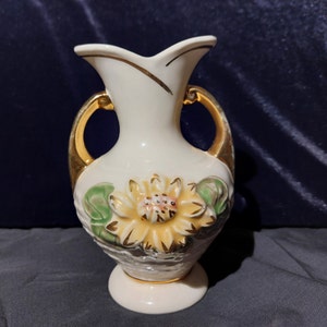 May include: A white ceramic vase with gold accents and a floral design. The vase has two handles and a wide mouth.