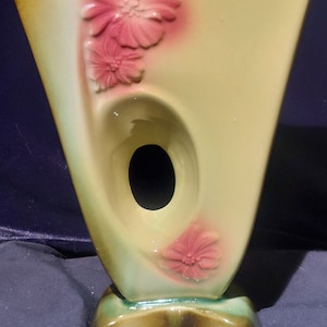 Puede incluir: Un jarrón de cerámica verde y marrón con una forma abstracta única. El jarrón tiene un corte en el medio y está decorado con flores rosas.