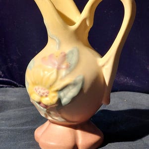 Könnte beinhalten: Eine gelbe Keramikkanne mit Blumenmuster. Die Kanne hat einen rosa Sockel und einen Henkel. Die Blumen sind gelb, rosa und grün.