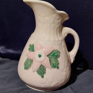 Könnte beinhalten: Eine antike Keramikkanne mit einem rosa-grünen Blumendesign. Die Kanne hat eine breite Öffnung und einen Henkel. Sie ist hellrosa mit einer weißen Glasur.
