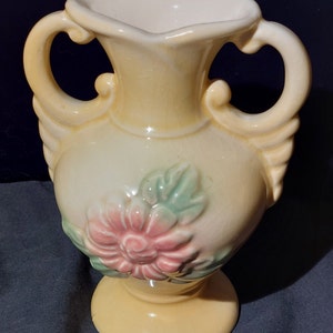 Puede incluir: Un jarrón de cerámica amarillo con dos asas y un diseño floral. El jarrón tiene una flor rosa con hojas verdes.