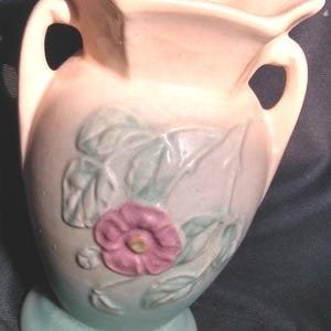 Peut inclure: Un vase en céramique vintage à deux anses, avec un motif floral composé d'une fleur rose et de feuilles vertes. Le vase est recouvert d'un glacis rose clair et bleu.
