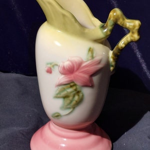 May include: A vintage ceramic pitcher with a pink base, a green handle, and a floral design featuring a pink flower with green leaves.