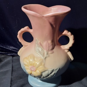 Puede incluir: Un jarrón de cerámica vintage con un esmalte rosa y azul. El jarrón tiene dos asas y está decorado con un diseño floral en tonos amarillos, rosas y verdes.