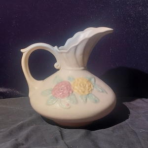 Pode incluir: Um jarro de cerâmica branca com um design floral. O jarro tem uma boca larga e uma alça. As flores são rosa, amarelas e verdes.