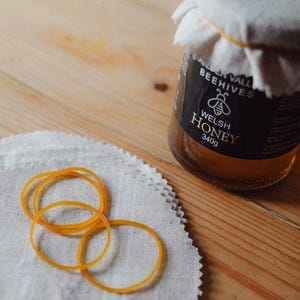 May include: A jar of Welsh honey with a black label and a white cloth cover, secured by a yellow rubber band. The jar sits on a wooden surface next to a stack of white fabric circles and several orange rubber bands.