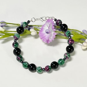 May include: A beaded bracelet featuring a mix of black, green, and purple beads. The bracelet includes large, glossy black beads interspersed with smaller beads in shades of green and purple. A small, light purple flower is attached to the bracelet.