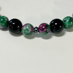 May include: A beaded bracelet featuring a combination of black, green, and ruby-colored beads. The bracelet's design includes polished black beads interspersed with green and ruby-colored stone beads, connected by a small, dark metal bead.