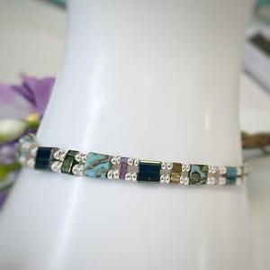 May include: A beaded bracelet featuring a repeating pattern of small, round, metallic silver beads interspersed with rectangular, flat-edged beads in shades of turquoise, dark blue, green, purple, and light pink, arranged in a single continuous strand.