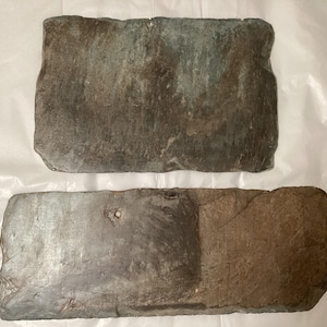 May include: Two rectangular stone slabs with uneven, rough edges and weathered surfaces. The top slab is a mottled gray-green, while the bottom slab features a combination of dark gray and earthy brown tones with a small, circular hole near the center.