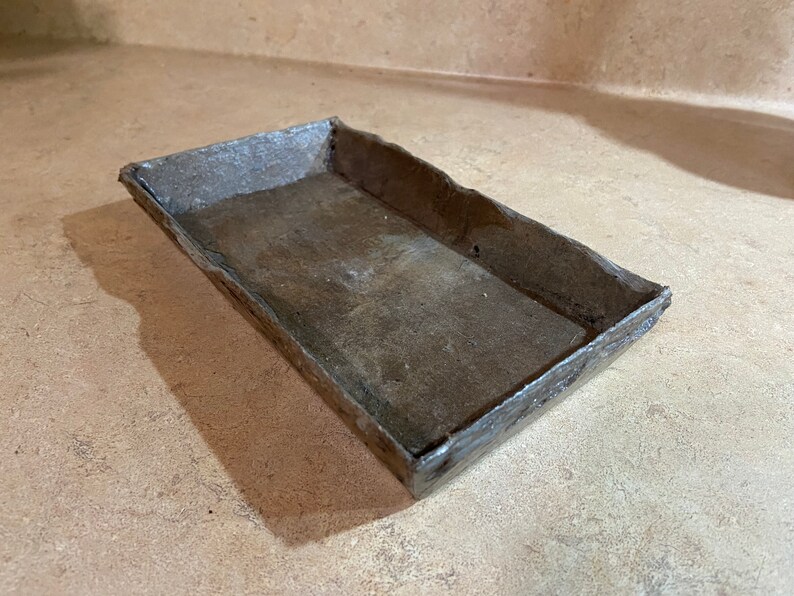 May include: A rectangular metal tray with a rustic, weathered finish. The tray has a slightly raised edge and a smooth, flat interior.