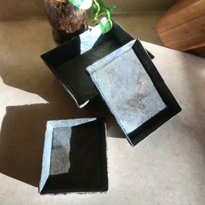 May include: Three square slate coasters with a dark gray color and a black border.