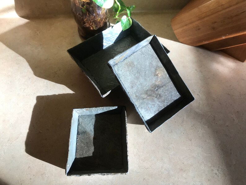 May include: Three square slate coasters with a dark gray color and a black border.