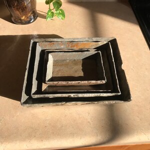 May include: Set of four nesting rectangular trays made of natural slate stone. The trays are in shades of gray, brown, and black. The trays are stacked on top of each other, with the largest tray at the bottom and the smallest tray at the top.