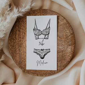 Könnte beinhalten: Eine schwarz-weiße Strichzeichnung eines BH- und Unterwäsche-Sets auf einer weißen Karte. Der BH ist im Bustier-Stil mit Spitzendetails. Die Unterwäsche ist im passenden Stil mit einer Schleife. Auf der Karte steht "36B" und "Medium". Die Karte liegt auf einem Holzbrett.
