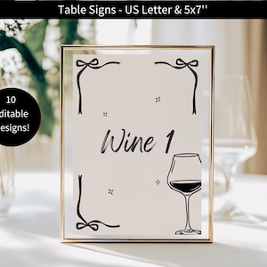 May include: A framed table sign with the text "Wine 1" and a wine glass illustration. The sign is in a gold frame and has decorative bow and star accents. The top of the sign reads "Table Signs - US Letter & 5x7" and a black circle with the text "10 Editable Designs!"