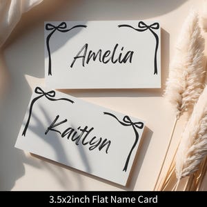 May include: Two white name cards with black calligraphy names, "Amelia" and "Kaitlyn". Each card has a black bow and ribbon design in the corners. The cards are 3.5 x 2 inches.