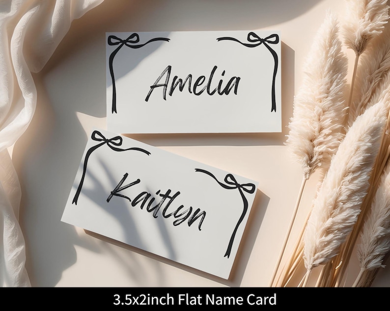 May include: Two white name cards with black calligraphy names, "Amelia" and "Kaitlyn". Each card has a black bow and ribbon design in the corners. The cards are 3.5 x 2 inches.