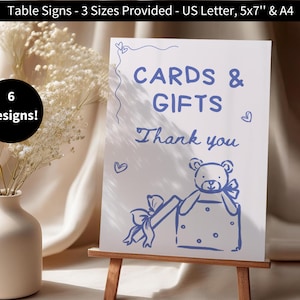 May include: A white sign with blue text that reads "CARDS & GIFTS Thank you". A blue teddy bear with a blue bow is holding a gift box. The sign is on a wooden easel.