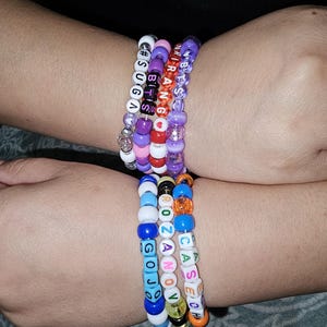 May include: A collection of six handmade beaded bracelets featuring colorful plastic pony beads and letter cubes. The bracelets display various text including #SUGA, BTS, SARIRANG, ROZANOV, GOJO, and CASECH, interspersed with small heart-shaped beads.