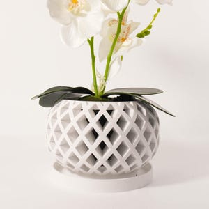 Può includere: Un vaso di ceramica bianco a forma di diamante con un'orchidea artificiale. L'orchidea presenta petali bianchi con accenti gialli e arancioni, steli verdi e foglie verde scuro. Il vaso poggia su un piattino bianco abbinato.