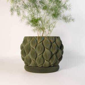 Op de afbeelding: Een decoratieve olijfgroene plantenpot met een textuur van bladeren. De pot staat op een bijpassende schotel en bevat een groene plant met dunne, delicate bladeren. De pot is rond en heeft een modern design.