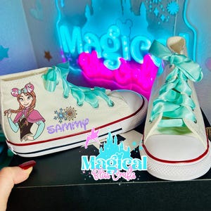 Può includere: Scarpe da ginnastica Converse alte bianche con opere d'arte personalizzate di Anna di Frozen e lacci verde menta. Le scarpe sono esposte su una scatola Converse nera con la scritta "CONVERSE" in bianco. Lo sfondo include un'insegna al neon con la scritta "Magic".