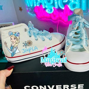 Può includere: Scarpe da ginnastica Converse alte bianche con lacci a nastro azzurro chiaro. Una scarpa presenta un'immagine dei cartoni animati di Elsa di Frozen, il nome "Jayla" e un disegno a fiocco di neve. Le scarpe sono su una scatola Converse nera.