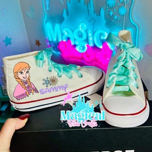 Può includere: Scarpe da ginnastica Converse alte bianche con lacci turchese. Una scarpa presenta un'immagine a cartoni animati di una principessa e il nome "Sammy". Le scarpe sono su una scatola Converse nera con la scritta "CONVERSE".