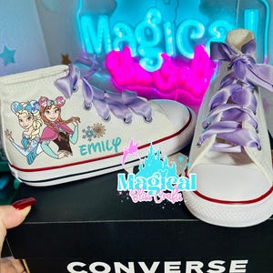 Può includere: Scarpe da ginnastica alte in tela bianca con lacci in nastro di raso viola. Le scarpe da ginnastica presentano una grafica a cartoni animati di due personaggi e il nome "Emily". Le scarpe sono su una scatola Converse nera.