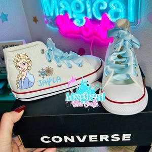 Può includere: Scarpe da ginnastica Converse alte bianche con lacci a nastro azzurro chiaro. Una scarpa presenta un'immagine dei cartoni animati di Elsa di Frozen, il nome "Jayla" e un disegno a fiocco di neve. Le scarpe sono su una scatola Converse nera.