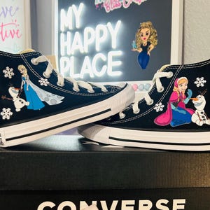 Può includere: Scarpe da ginnastica Converse alte nere con illustrazioni di Elsa e Olaf di Frozen, con lacci e suole bianche. Le scarpe sono su una scatola Converse nera. Lo sfondo include un cartello con la scritta "My Happy Place".