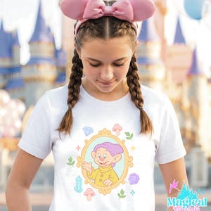 May include: White t-shirt featuring a cartoon image of Dopey from Snow White inside a gold frame, surrounded by colorful gems and mushrooms. The shirt is worn with pink bow ears. The text "Magical Blue Castle" is visible on the right side.