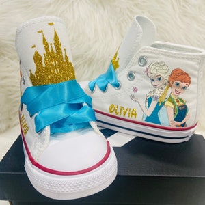 Può includere: Scarpe Converse bianche alte con un nastro blu e un design di castello con glitter dorati. L'altra scarpa ha un design Frozen con Elsa e Anna. Il nome "Olivia" è scritto sul lato della scarpa.