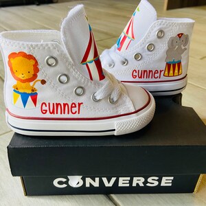 K&ouml;nnte beinhalten: Wei&szlig;e Converse-Sneaker mit hohem Schaft und einem roten, gelben und blauen Zirkuszeltdesign. Ein Schuh hat einen L&ouml;wen und der andere einen Elefanten. Der Name "Gunner" ist auf beiden Schuhen geschrieben.