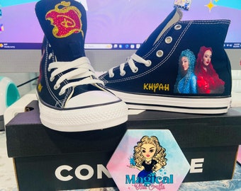 May include: Black high-top sneakers with white laces and soles. One shoe features a glittery design and the other has a portrait of two women and the name "Khyah" in gold. The shoes are on a black box with the word "CONVERSE".