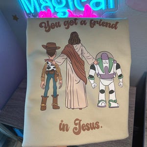 Puede incluir: Camiseta verde claro con una ilustraci&oacute;n de Jes&uacute;s, Woody y Buzz Lightyear. El texto "You got a friend in Jesus" est&aacute; impreso en marr&oacute;n. La camiseta tiene cuello redondo.