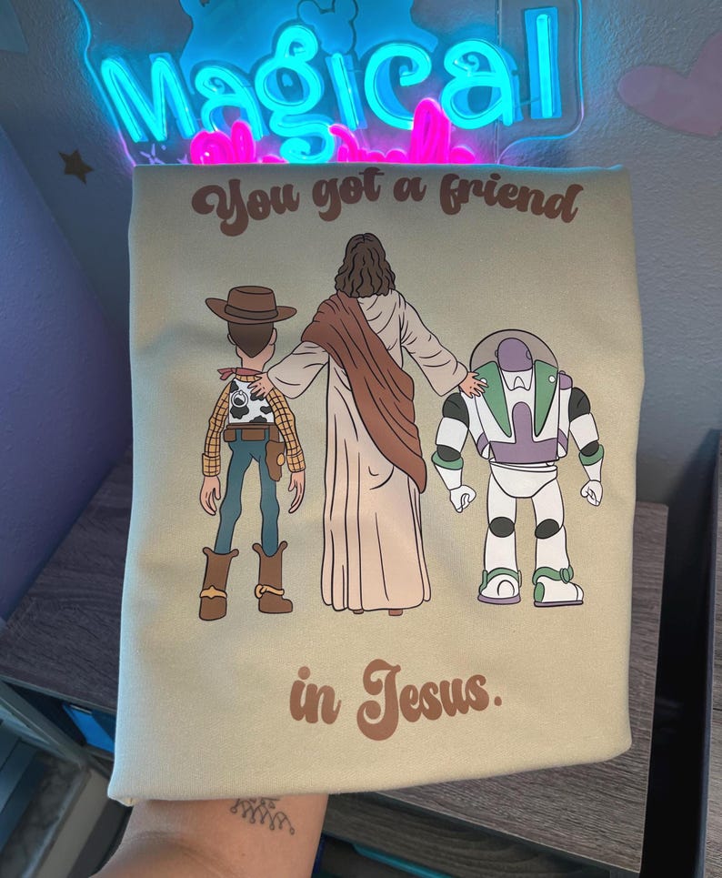 Puede incluir: Camiseta verde claro con una ilustraci&oacute;n de Jes&uacute;s, Woody y Buzz Lightyear. El texto "You got a friend in Jesus" est&aacute; impreso en marr&oacute;n. La camiseta tiene cuello redondo.