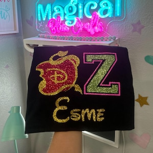 May include: Black t-shirt with a glittery red apple design and the letter "Z" in green with a pink outline. The name "Esme" is written in gold glitter below the letter. The shirt is likely a custom design.