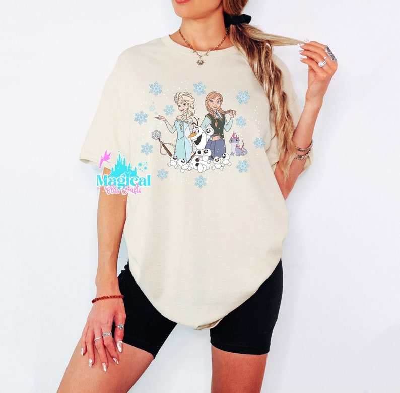 Op de afbeelding: Een cr&egrave;mekleurig T-shirt met een grafische print van Elsa en Anna uit Frozen. De afbeelding bevat ook Olaf en een kleine paarse draak. Het shirt wordt gedragen door een persoon.