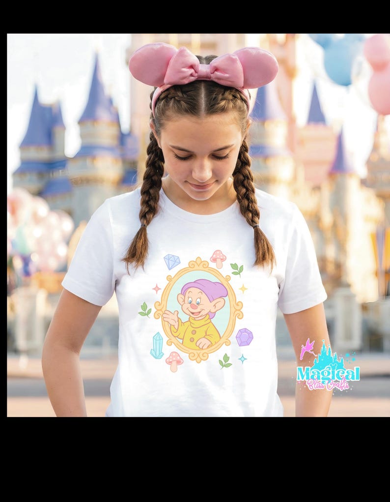 May include: White t-shirt featuring a cartoon image of Dopey from Snow White inside a gold frame, surrounded by colorful gems and mushrooms. The shirt is worn with pink bow ears. The text "Magical Blue Castle" is visible on the right side.