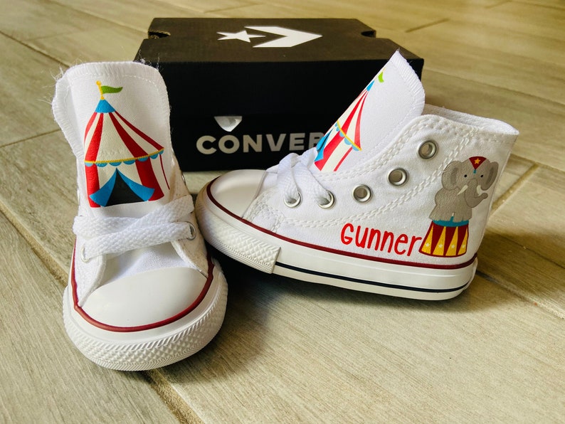 K&ouml;nnte beinhalten: Wei&szlig;e Converse-Sneaker mit hohem Schaft, auf einem Schuh ein rot-wei&szlig;-blaues Zirkuszeltdesign und auf dem anderen Schuh ein grauer Elefant mit einem rot-wei&szlig;-gelben Zirkuszeltdesign. Der Name "Gunner" ist in Rot auf dem Elefantenschuh geschrieben.