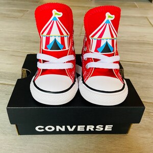 K&ouml;nnte beinhalten: Ein Paar rote Converse High-Top-Sneaker mit einem Zirkuszeltdesign auf der Vorderseite. Die Sneaker stehen auf einem schwarzen Converse-Schuhkarton.