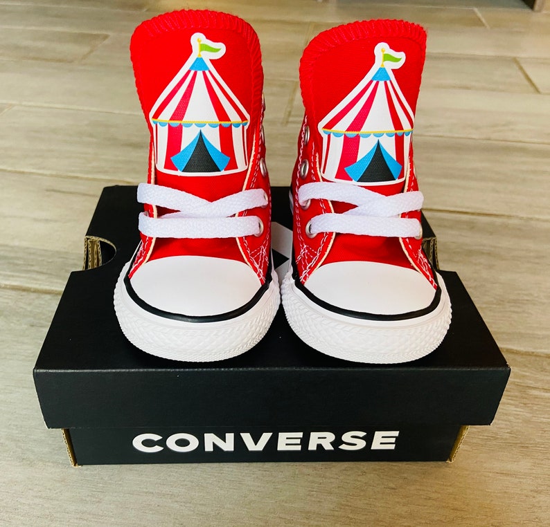 K&ouml;nnte beinhalten: Ein Paar rote Converse High-Top-Sneaker mit einem Zirkuszeltdesign auf der Vorderseite. Die Sneaker stehen auf einem schwarzen Converse-Schuhkarton.